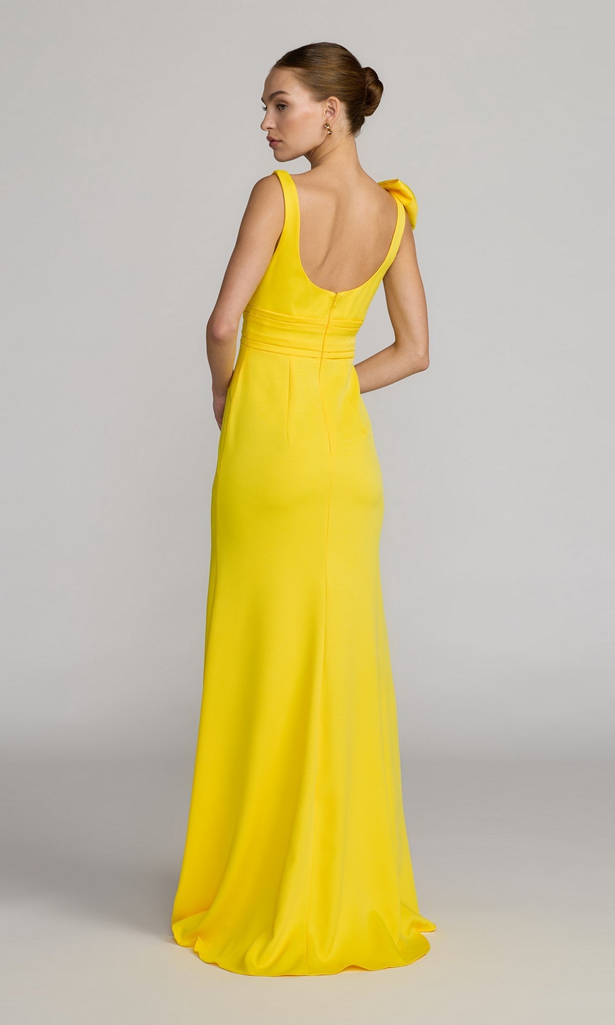 A woman stands with her back mostly to the camera, her hair styled in a neat bun, wearing the Long Formal Dress: Mac Duggal 49964—a floor-length, sleeveless yellow crepe column gown—set against a neutral background.